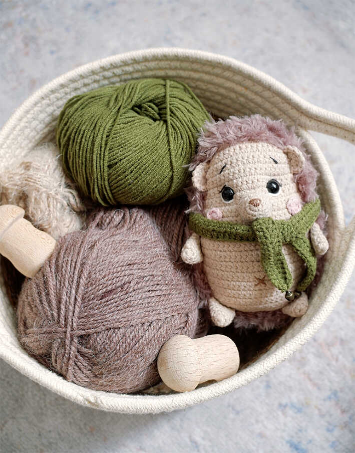Baby hedgehog amigurumi with fluffy spikes placed in basket with yarn skeins, handmade crochet project materials